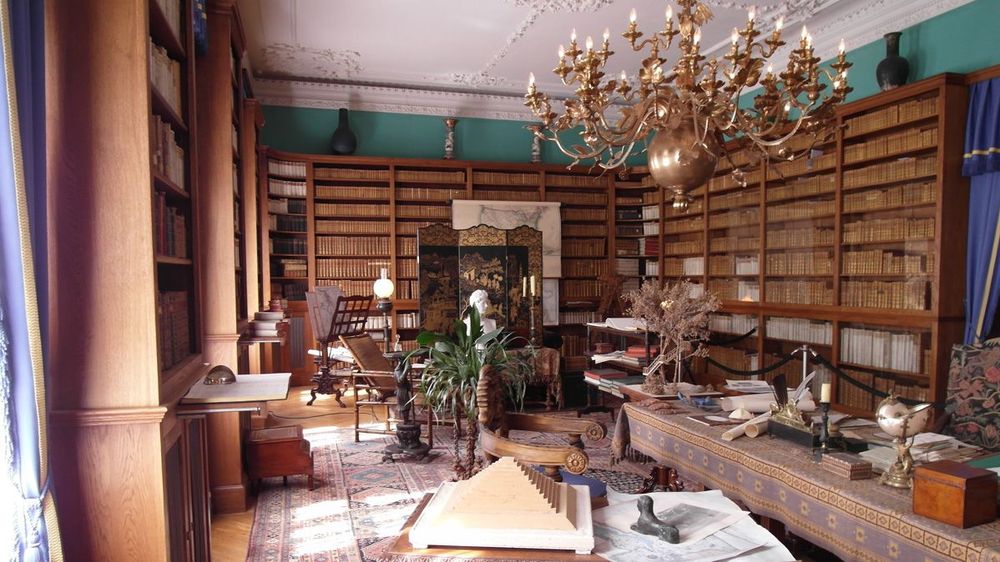 Innenansicht. Blick in die Bibliothek mit Bücherregalen, einem alten Schreibtisch und Sitzmöglichkeieten. An der Decke hängt einer großer vergoldeter Kronleuchter.