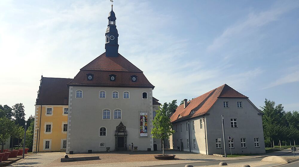 Ansicht auf einen Platz. Im Hintergrund steht ein hohes, weißes Haus mit rotem Sitzdach und Uhrenturm.
