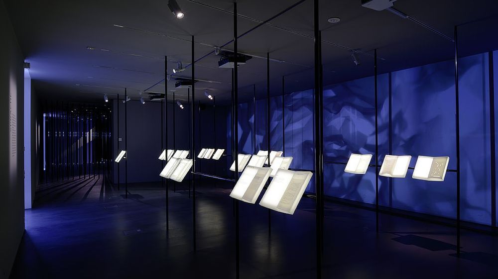 Innenansicht. Ausstellungsraum mit dunkelblauen Wänden. Vitrinen in Buchform liegen auf einem Metallgerüst.