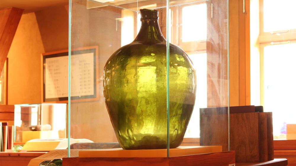 Innenansicht. Im Vordergrund steht eine Glasvitrine mit einer großen, dickbauchigen, grünen Flasche. Im Hintergrund zwei Fenster.