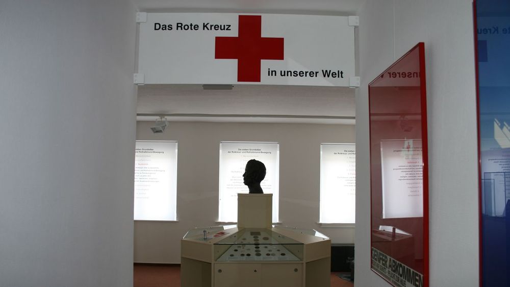 Inenansicht. Blick in einen Ausstellungsraum mit einer Ausstellungsvitrine und beleuchteten Texttafeln. An der Decke hängt ein Schild mit der Aufschrift "Das Rote Kreuz in unserer Welt". 