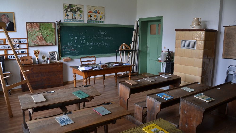 Innenansicht. Der Ausstellungsraum ist wie ein Klassenzimmer hergerichtet. Bänke, ein Lehrertisch, eine grüne Schiefertafel und Karten an den Wänden.