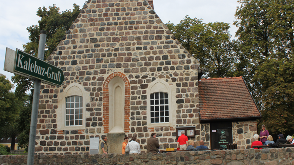 Außenansicht der Dorfkirche mit Menschen, die auf Bänkenb davor sitzen. Wegweiserschild mit der Aufschrift "Kalebuz-Gruft".