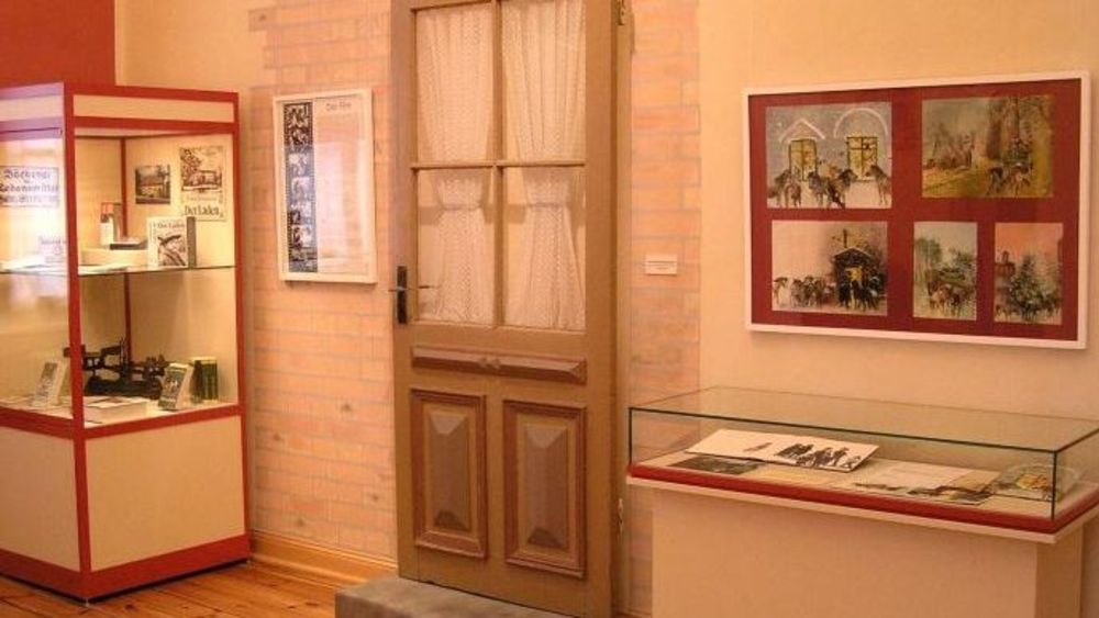 Innenansicht. Ausstellungsraum mit einer Hauseingangstür und zwei Glasvitrinen mit Ausstellungsobjekten. Informationstafeln und Bilder hängen an der Wand.