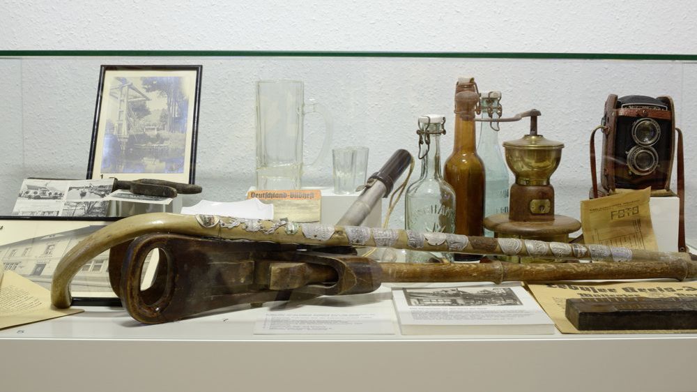 Innenansicht. Blick in eine Vitrine. Ausgestellt sind alte Flaschen, Gläser, ein Wanderstock, eine alte Kamera und verschiedene Dokumente. 