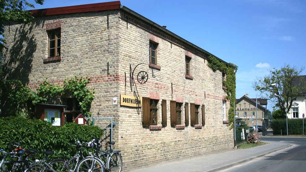 Außenansicht auf ein aus Zieglen erriAuf einer Längswand steht "Dorfmuseum". Vor dem Haus stehen Fahrräder.chtetes Haus. 