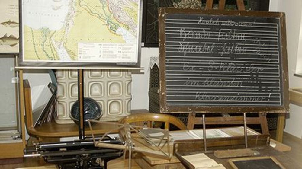 Innenansicht in einen Ausstellungsraum in der Art eines Klassenzimmers: Schulbank, Schreibtafel, Kartenmaterial. 