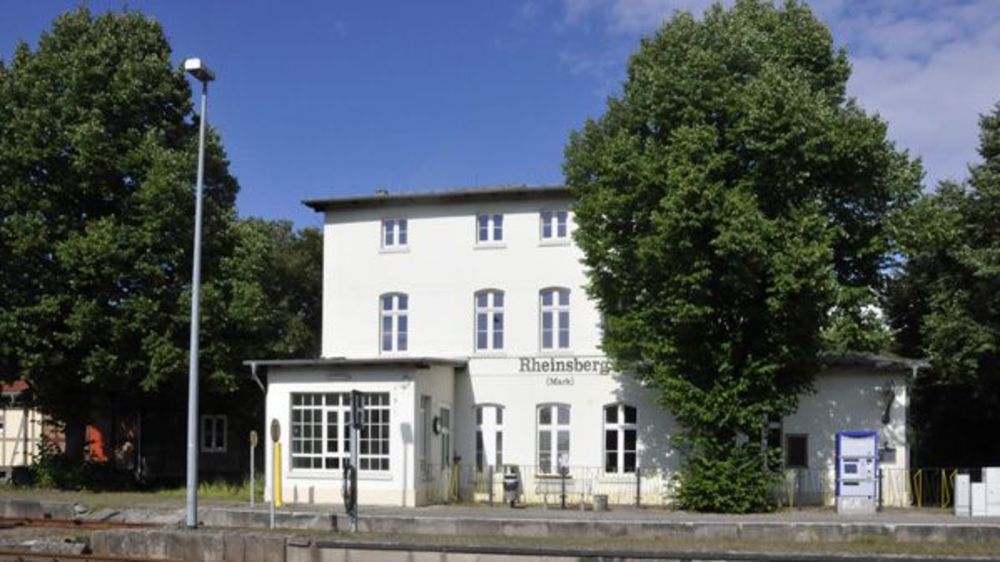 Außenansicht. Blick auf einen alten, restaurierten, Bahnhof, weiß getüncht. Auf der Fassade steht "Rheinsberg (Mark)". 