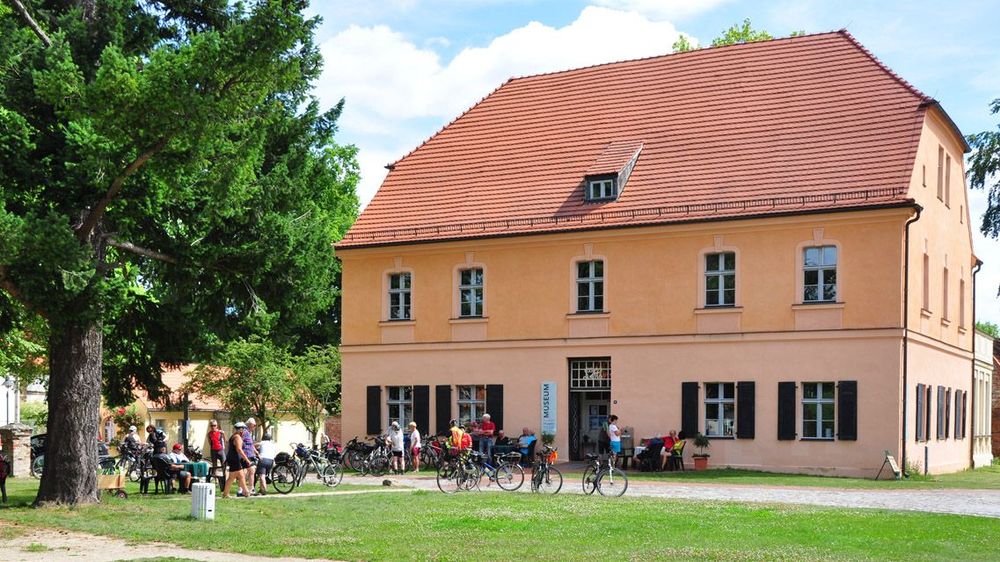 Außenansicht eines dreistöockigen alten Hauses mit hoch anstehendem Dach. Vor dem Eingang befindet sich eine große Menschengruppe mit Fahrrädern.