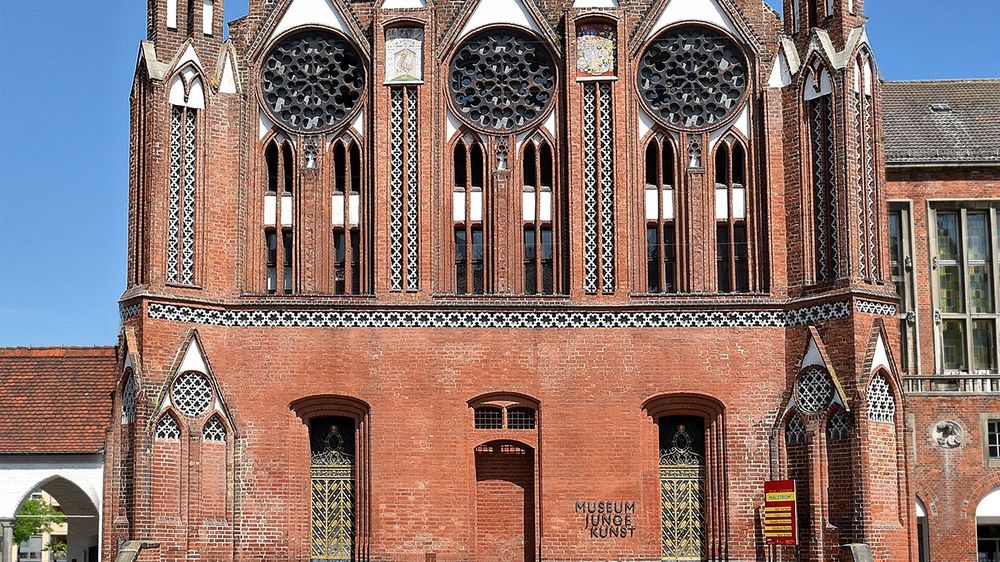 Außenansicht. Vorderansicht der Rathaushalle. Ein roter Klinkerbau mit gotischen Fenstern und Rosettenfenstern.